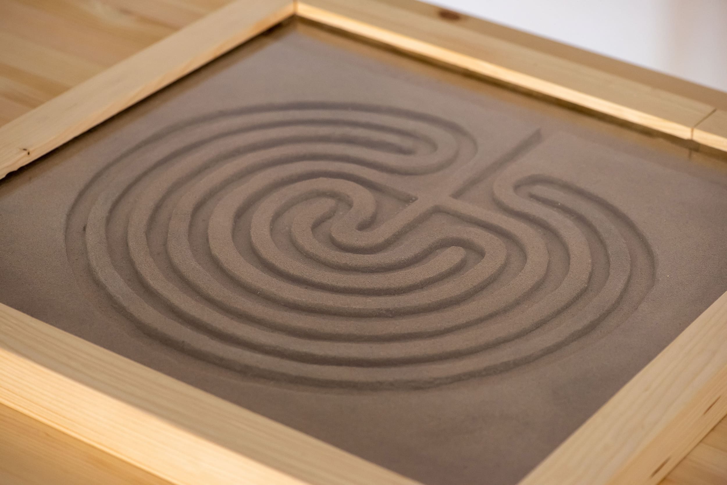 A wooden-framed sand garden with a spiral Cretan labyrinth pattern. The smooth sand and curved lines create a calming and meditative atmosphere.