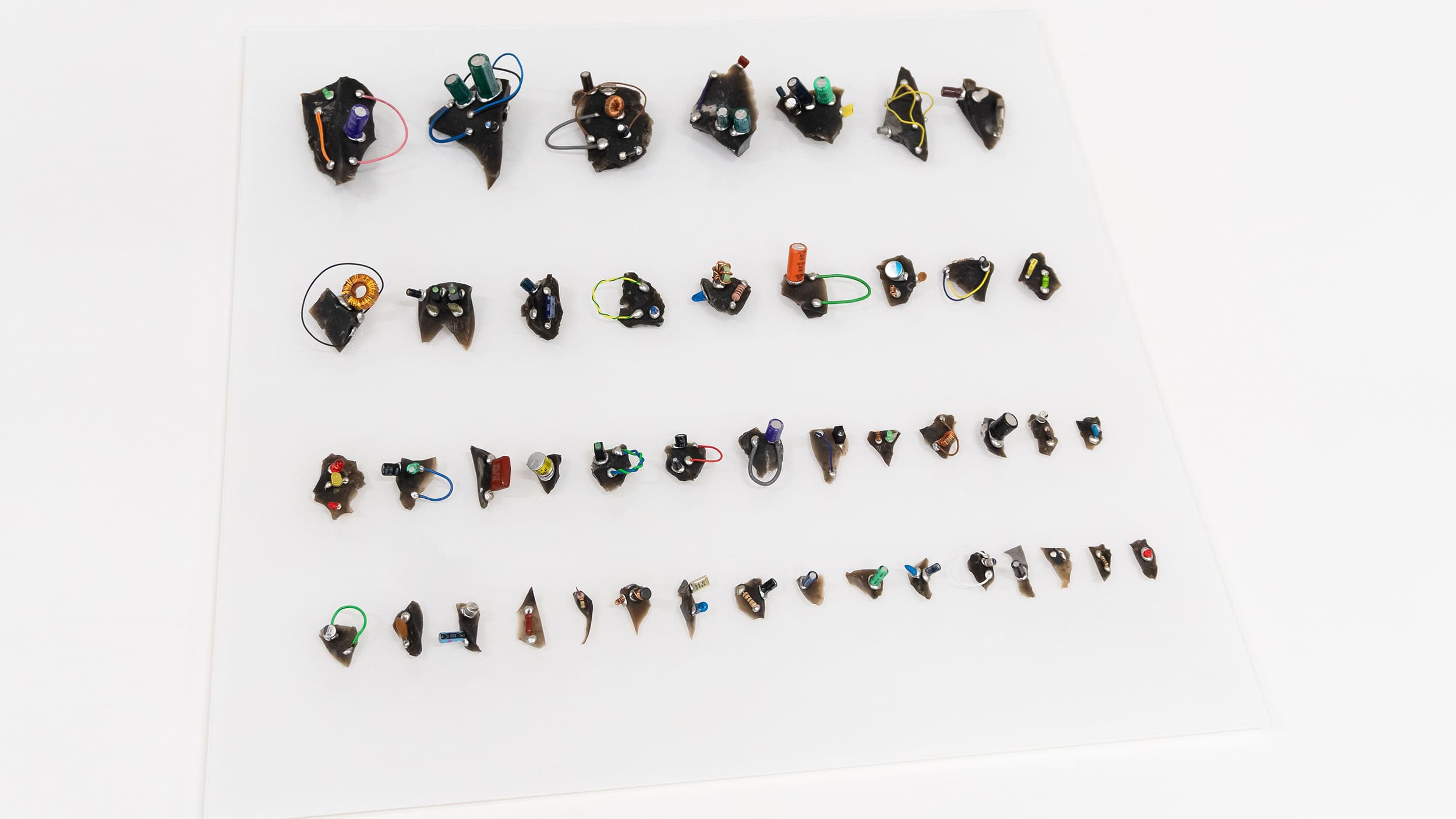 Array of small, abstract sculptures resembling circuit boards, with colorful wires and components, neatly aligned on a white background.