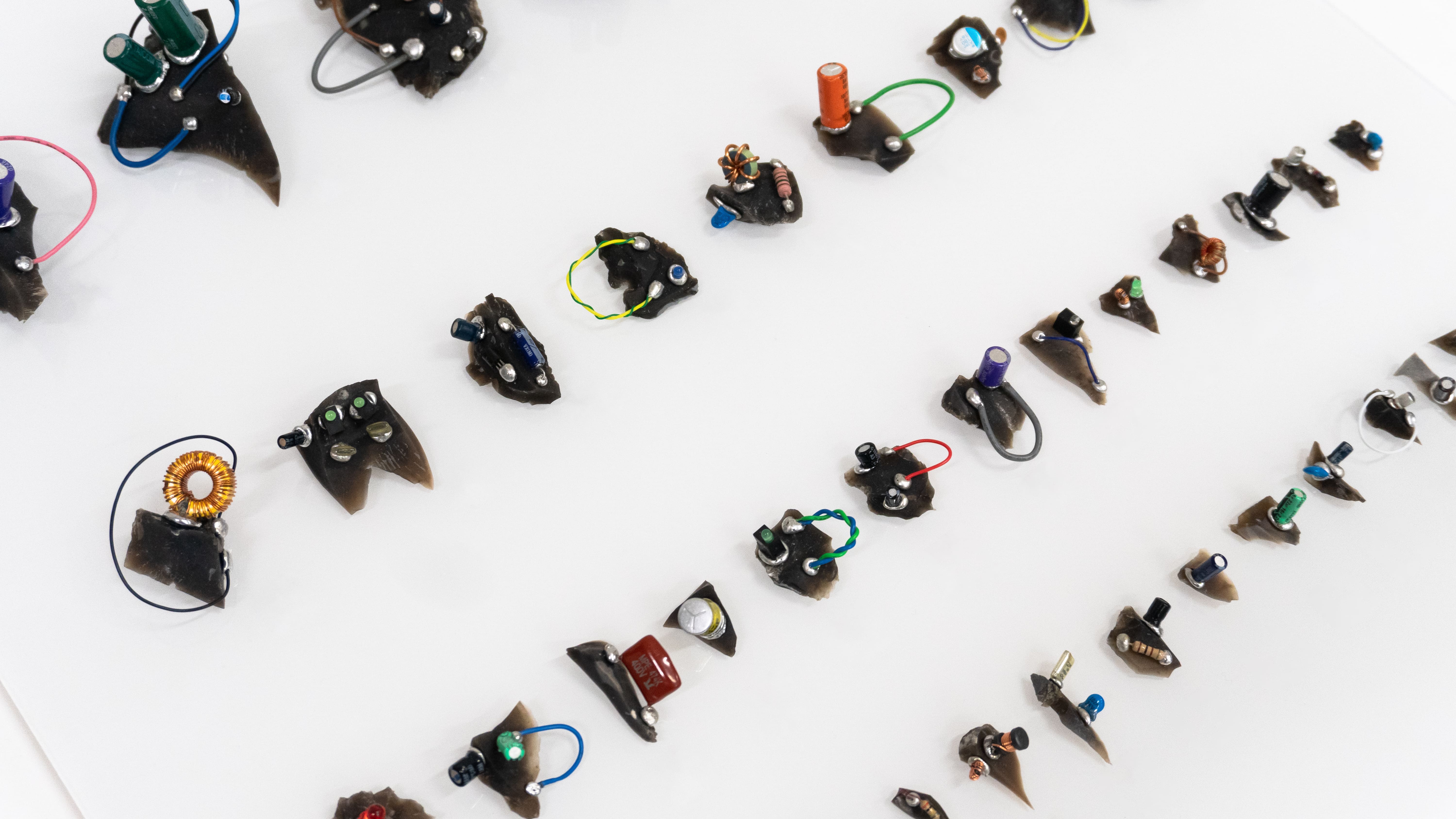Array of small dark sculptures on a white surface, each adorned with colorful wires and electronic components, creating a playful, inventive atmosphere.