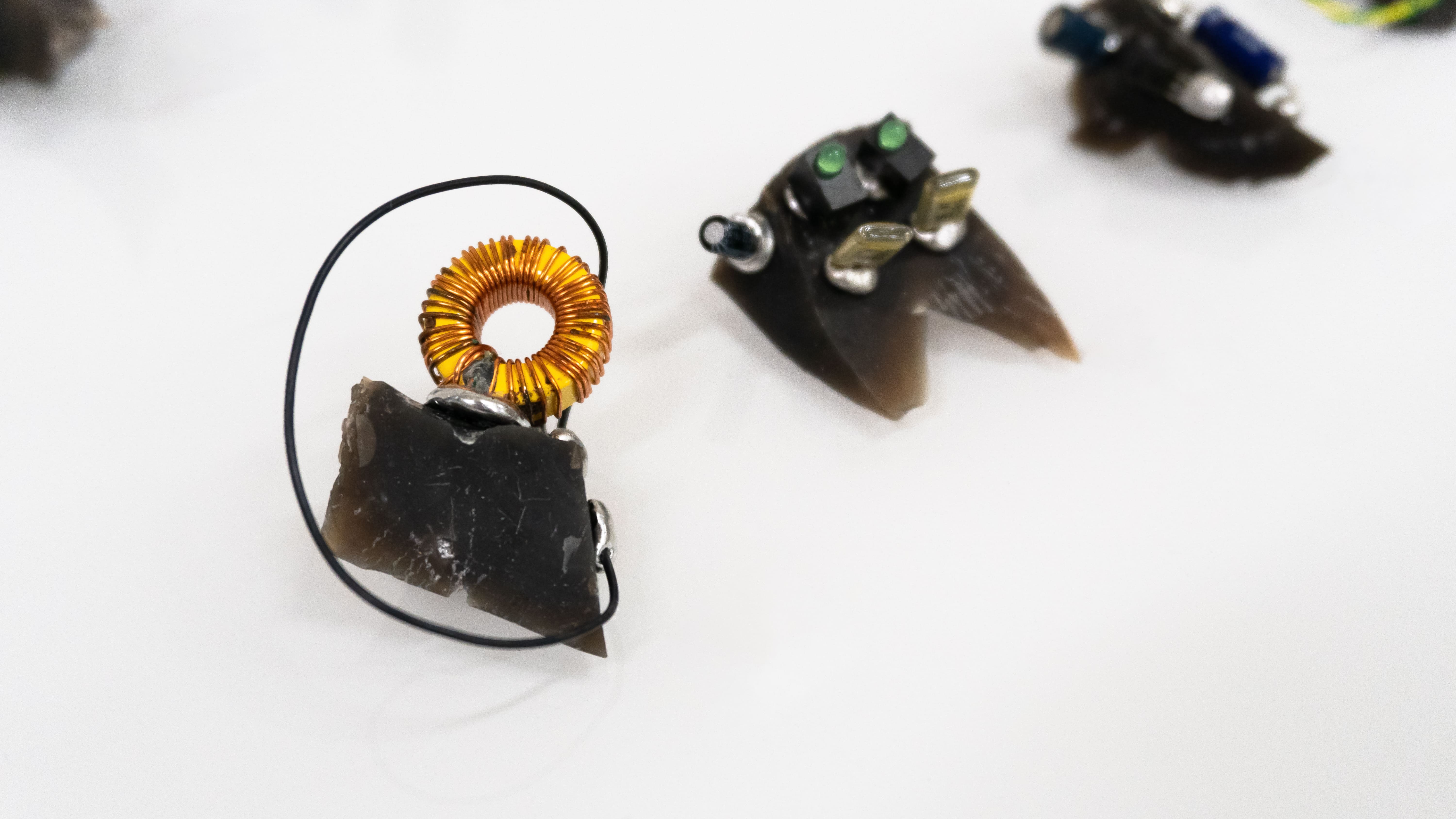 Close-up of small electronic sculptures on a white background. Features a copper coil on a stone and various components, conveying a futuristic, artistic vibe.