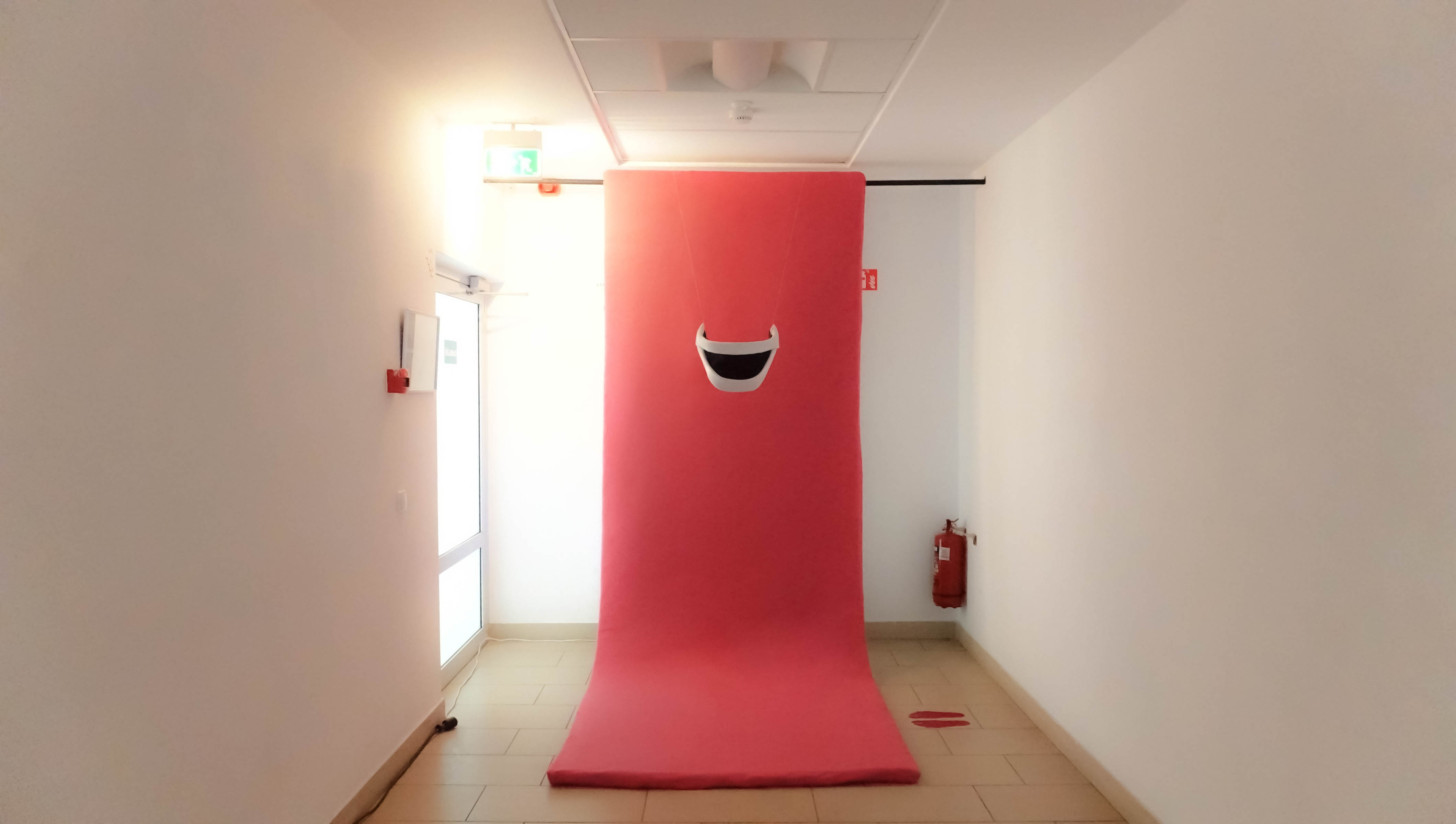 A hallway with white walls features a large pink mattress backdrop, displaying a white smiling mouth in the middle. A fire extinguisher sits on the right.