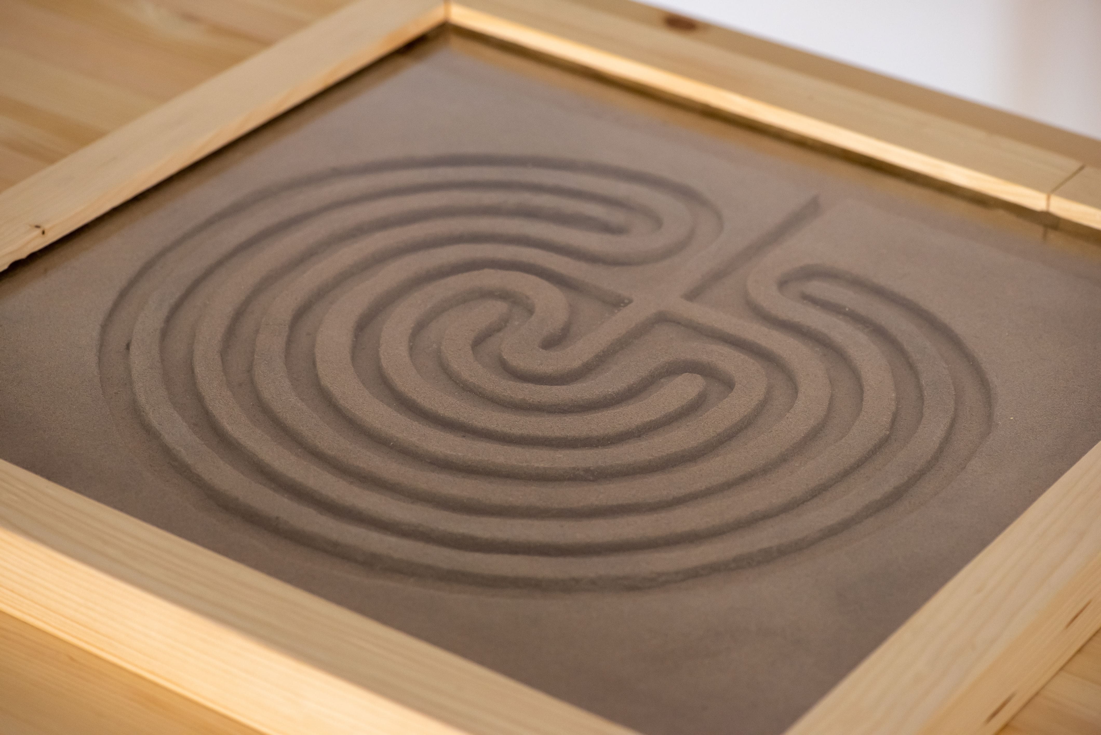 A wooden-framed sand garden with a spiral Cretan labyrinth pattern. The smooth sand and curved lines create a calming and meditative atmosphere.